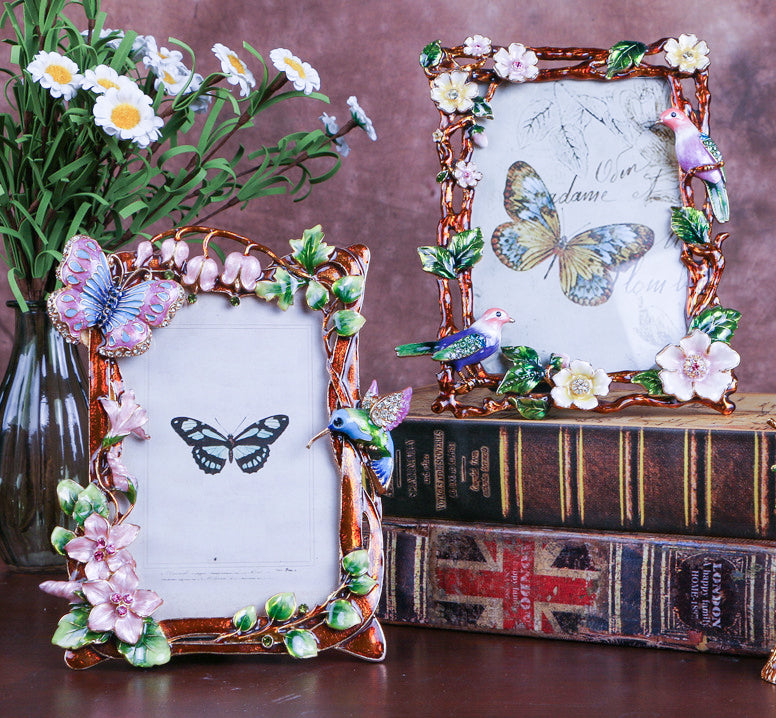 Two exquisite enamel photo frames with intricate, hand-painted floral and butterfly designs. One features a butterfly and pink flowers, while the other highlights birds and blossoms. Perfect for adding an artistic and vintage touch to home decor.